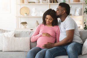 Photo of a Black couple sitting on the couch together while the man kisses his partner's head. The pregnant woman in this photo illustrates the many women who would benefit from pregnancy and prenatal counseling in Chicago, IL. 60604 | 60605 | 60607