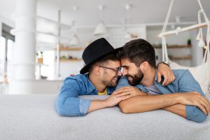 Image of a male same-sex couple sitting on the couch and smiling in an embrace. This image represents the happiness couples can find when working with a PMH-C for third party reproduction in Chicago, IL. 60305 | 60130 | 60402